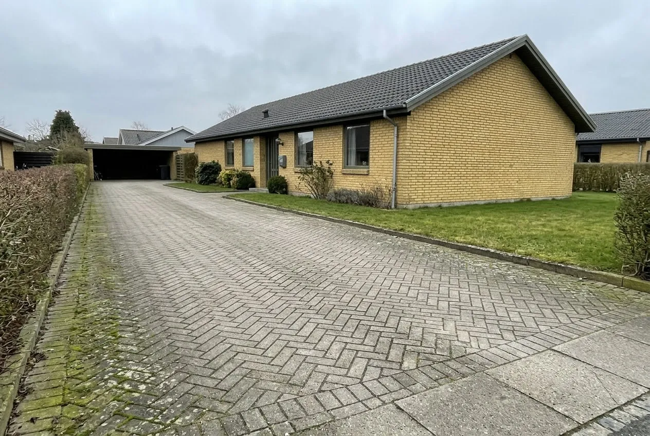 Gult murstenshus med indkørsel af grå belægningssten i sildebensmønster, hvor algerne er begyndt at sætte sig